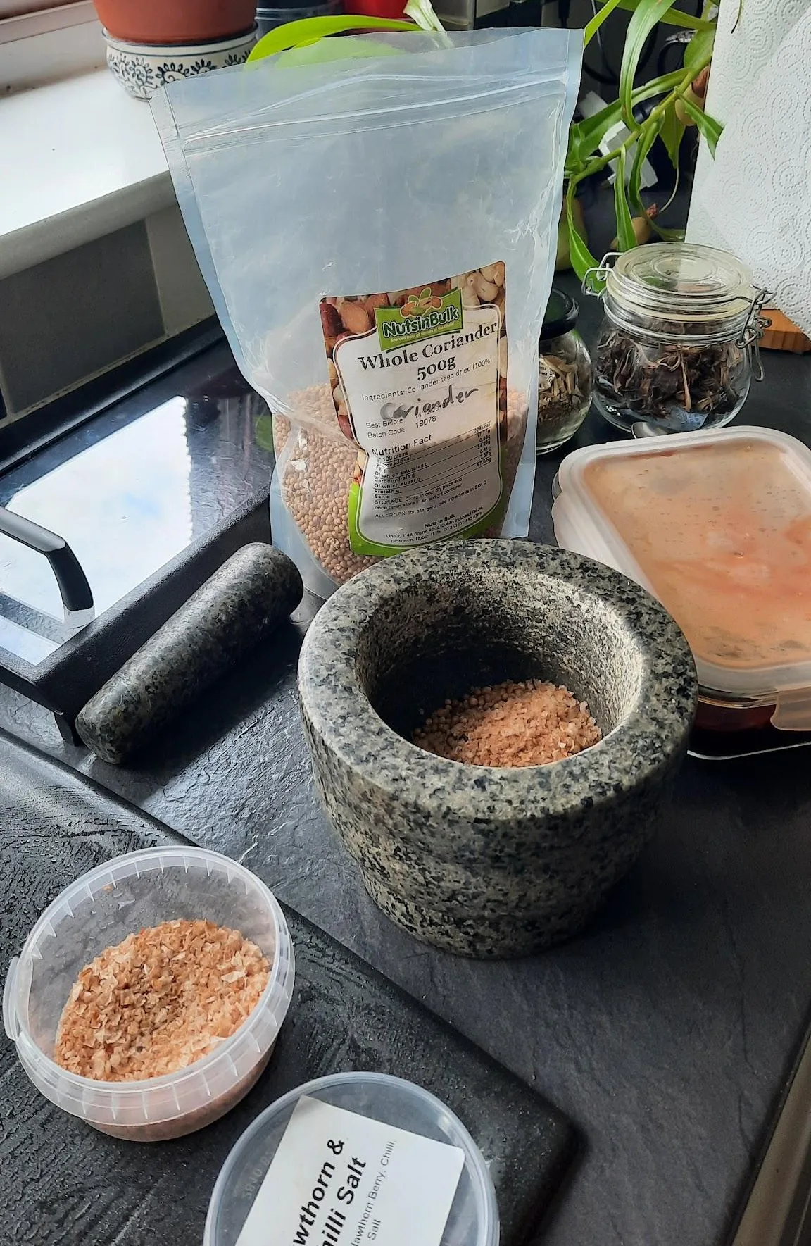 A granite pestle and mortar with coarse salt and coriander seed, beside bags and bowls of the spice mix